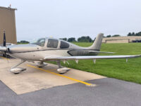 Silver Cirrus SR22 single-engine airplane parked on tarmac at small airport, general aviation aircraft side view.