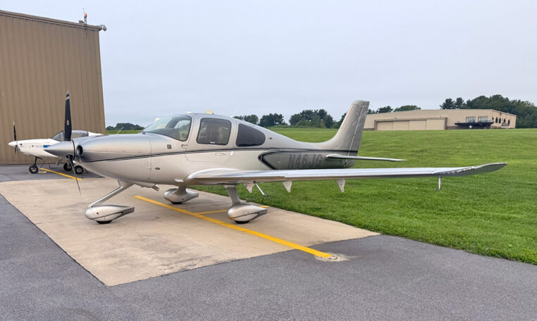 Silver Cirrus SR22 single-engine airplane parked on tarmac at small airport, general aviation aircraft side view.