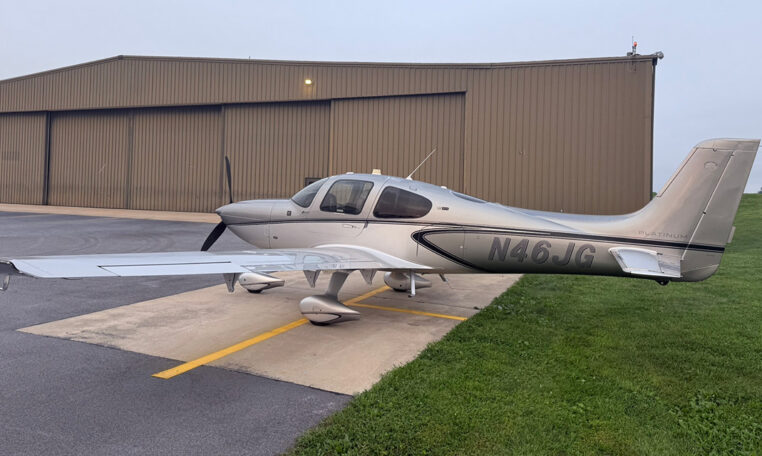 Silver Cirrus SR22 single-engine propeller plane N46JG parked on airport ramp beside hangar.