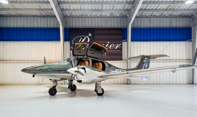 Twin-engine light aircraft with gullwing doors open in a hangar, showcasing tan leather interior and sleek silver finish.